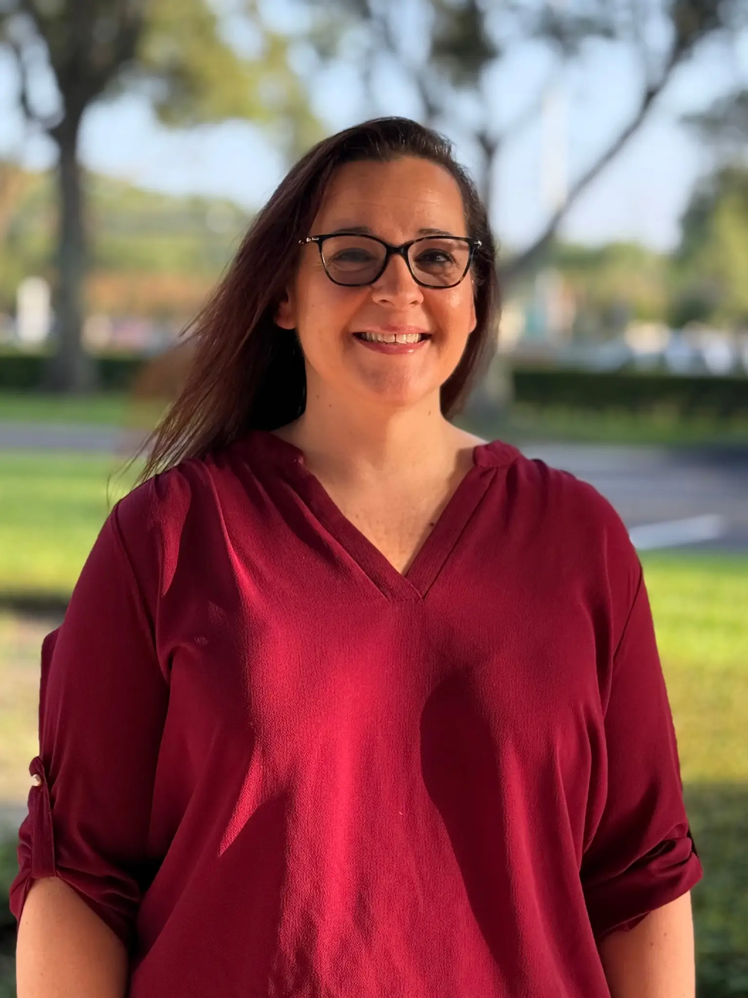 Smiling woman with glasses and long dark hair wearing a maroon V-neck top outdoors in a park-like background.