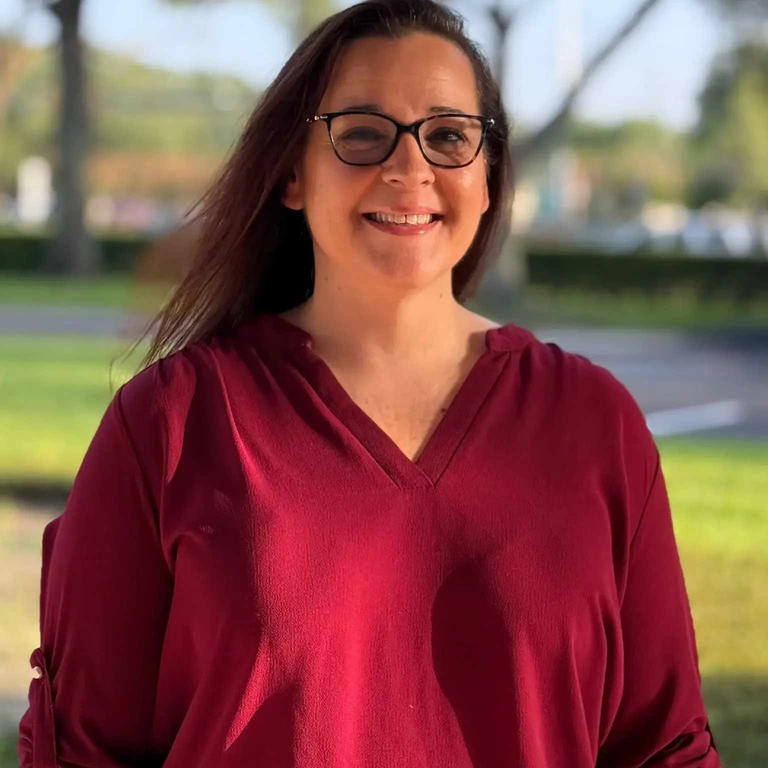 Smiling woman with glasses and long dark hair wearing a maroon V-neck top outdoors in a park-like background.